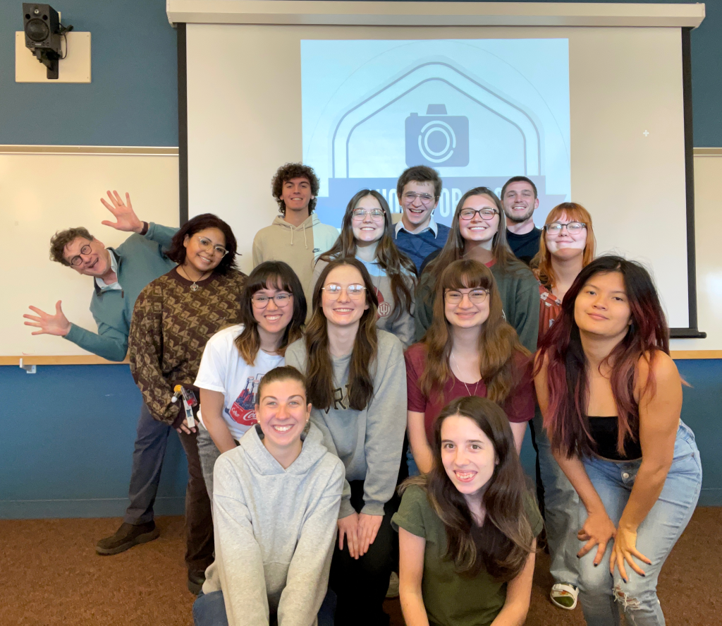 A group of students and professionals posing for a photo in a classroom setting, with a presentation screen in the background displaying a camera logo.